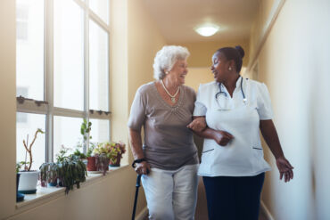 Nurse assisting elderly woman in hallway.